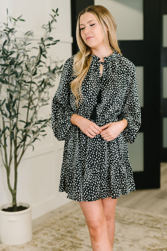 Woman wearing a black and white patterned dress standing indoors with a plant in the background