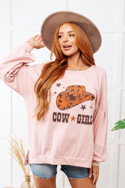 Woman wearing a pink sweatshirt with a cowgirl graphic and text, standing against a white wall.