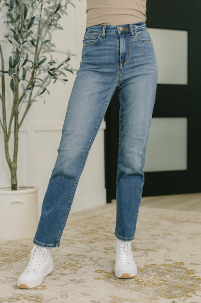 Person wearing blue jeans and white sneakers standing indoors with a plant in the background