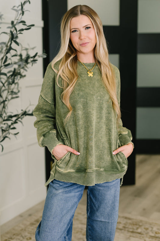 Woman wearing a green sweatshirt and blue jeans standing indoors with a plant in the background