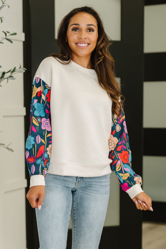 Woman wearing a cream sweater with colorful floral sleeves and light blue jeans.