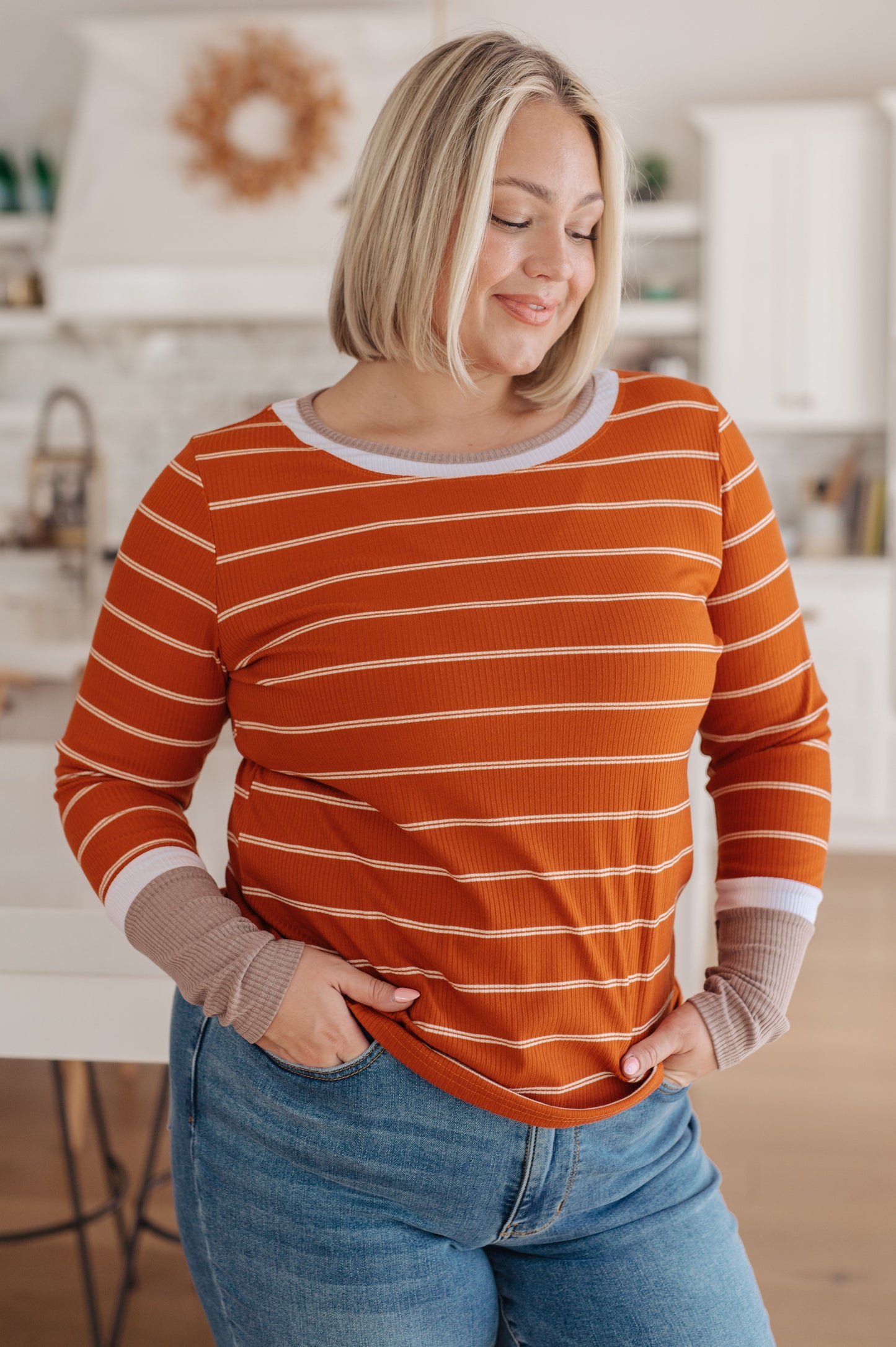 Woman wearing an orange striped sweater and blue jeans in a kitchen.