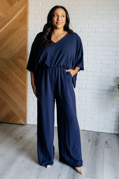 Woman wearing a navy blue jumpsuit standing against a white brick wall.