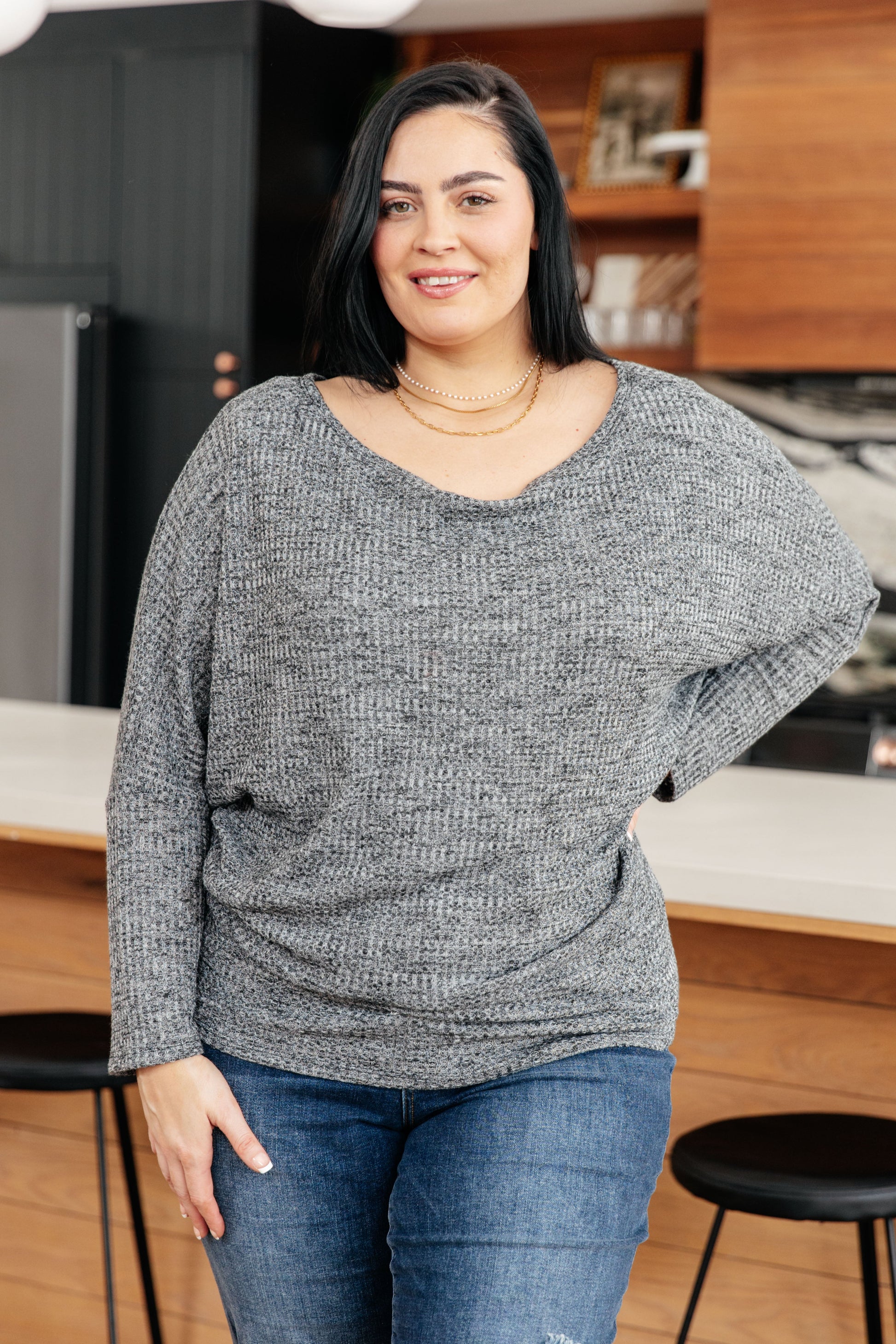 Woman wearing a gray sweater and blue jeans standing in a kitchen.